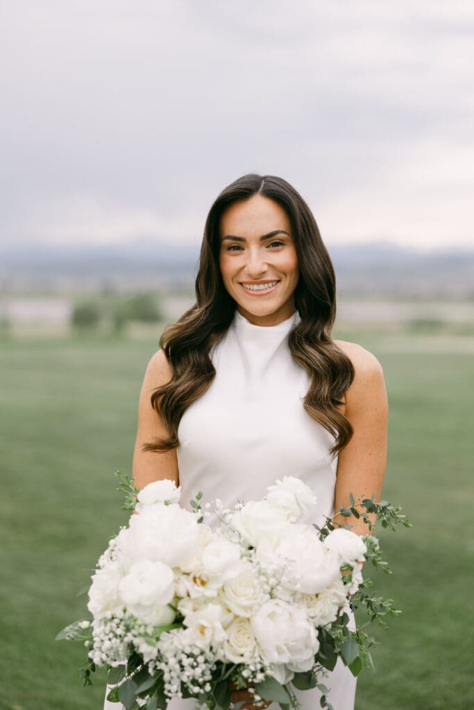 Bride smiling and holding an all-white bouquet on the green lawn before the ceremony
