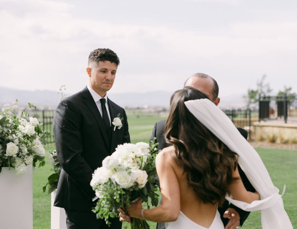 Bride with a flowing veil and white bouquet meets her groom at the altar