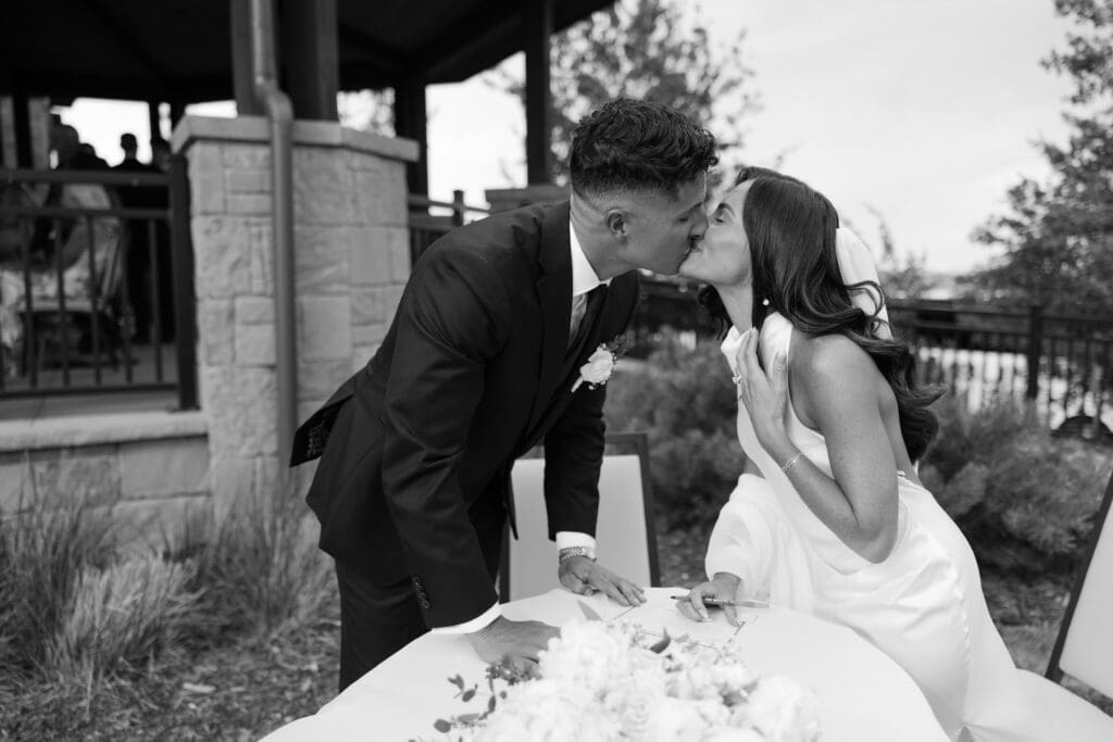 Bride and groom lean across the table for a kiss on the clubhouse patio in black and white