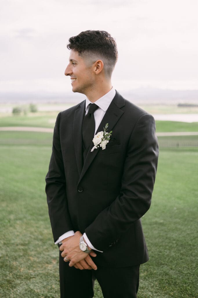 Groom in a black suit and white boutonniere standing on the golf course lawn