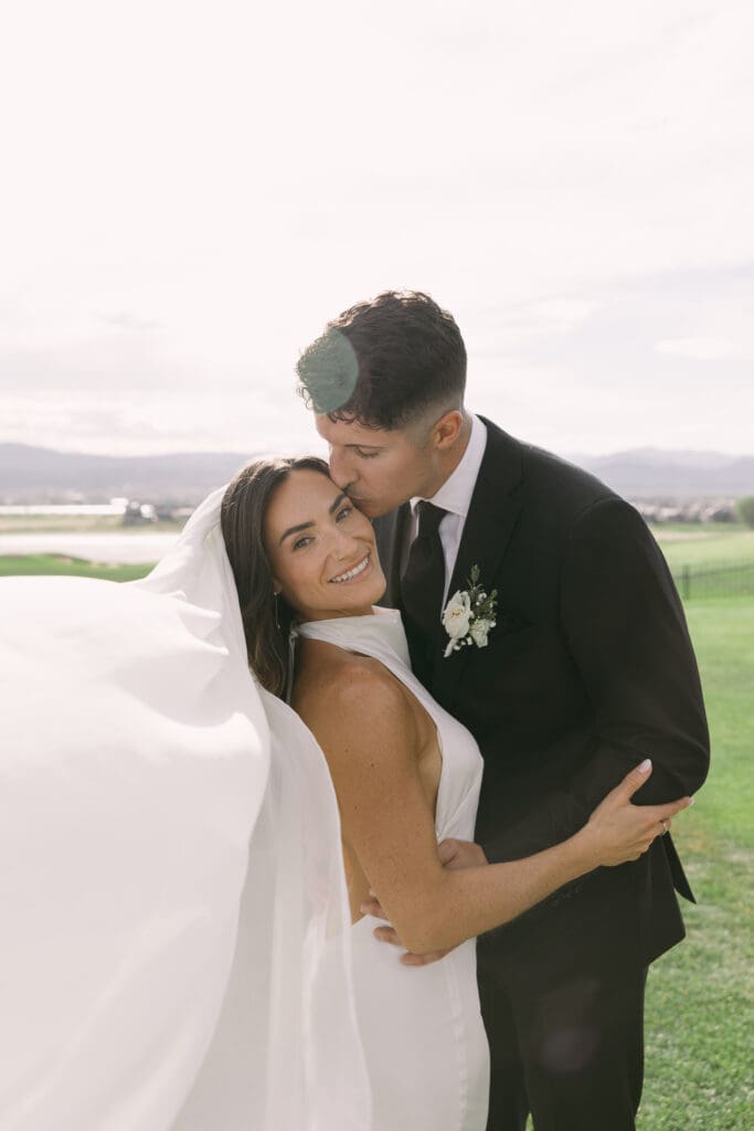Groom kisses his bride's cheek as her veil catches the breeze at TPC Colorado