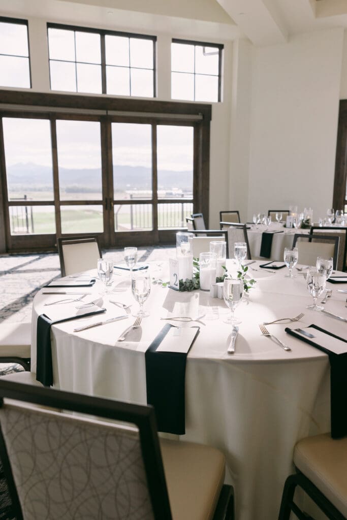 Reception tables set with white linens and black napkins beside tall windows at TPC Colorado