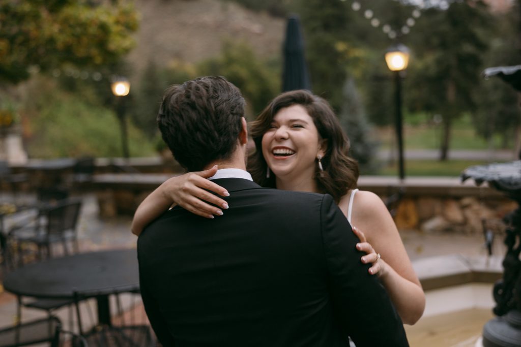 Bridgette laughs as Logan hugs her on the fountain patio with string lights and red rocks behind