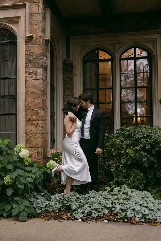 Bridgette rises on her toes to kiss Logan in front of the arched windows at Glen Eyrie Castle