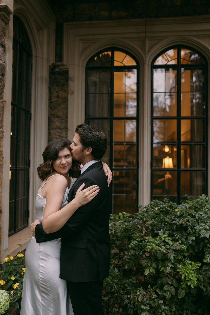 Couple hugging in front of tall arched windows glowing with warm light at Glen Eyrie Castle