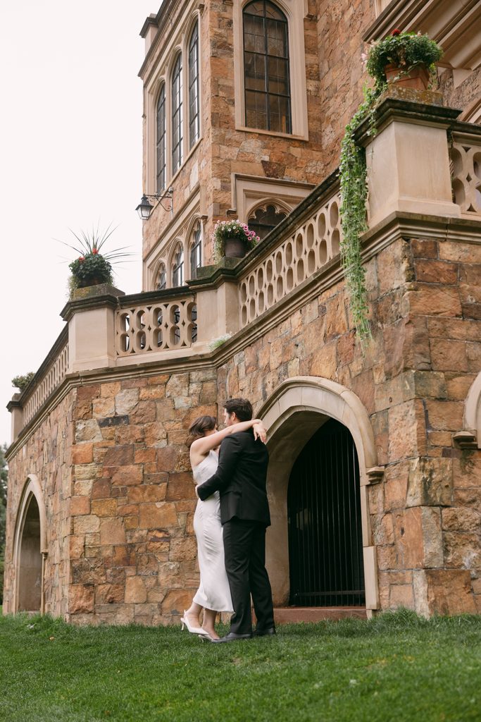 Couple embracing beneath a stone balcony and arched doorway at Glen Eyrie Castle
