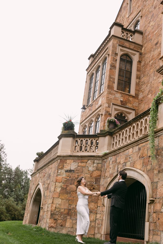 Bridgette and Logan dance hand in hand on the lawn beside the sandstone walls of Glen Eyrie Castle